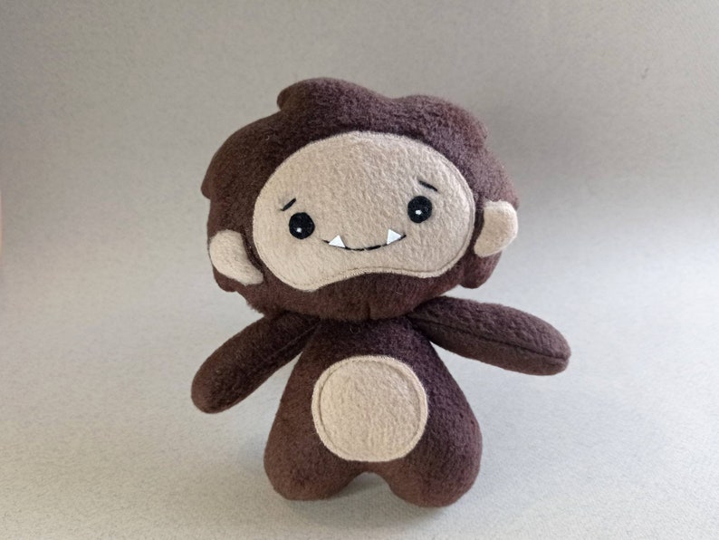 May include: A handmade brown and tan plush toy in the shape of a cartoon yeti or bigfoot. The plush has a round body, small arms, and a friendly face with a smiling mouth and two small teeth. The toy is made of soft fabric.