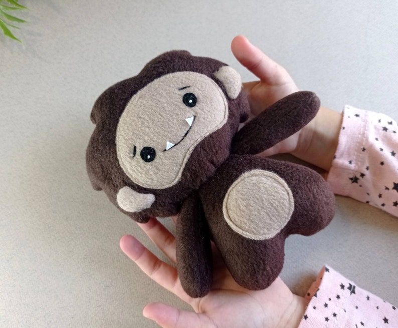 May include: A handmade brown felt Bigfoot plush toy with a tan face, arms, and feet. The Bigfoot has a smiling mouth, two white teeth, and black eyes. The plush toy is held in a person's hands.