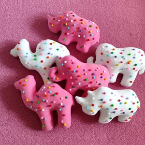 May include: A collection of plush animal toys, including a camel, elephant, and rhinoceros, in pink and white. Each toy is decorated with colorful, round sprinkles. The toys are arranged on a pink surface.