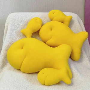 May include: Four yellow plush fish cushions of varying sizes. The cushions are arranged on a white fluffy surface.
