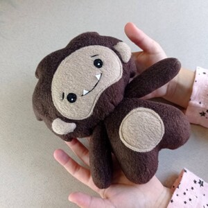 May include: A handmade brown felt Bigfoot plush toy with a tan face, arms, and feet. The Bigfoot has a smiling mouth, two white teeth, and black eyes. The plush toy is held in a person's hands.