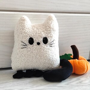 May include: White plush ghost cat with black embroidered eyes and whiskers, sitting next to a small orange plush pumpkin.