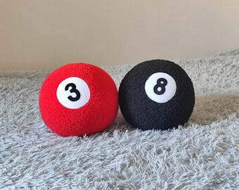 May include: Two plush spherical toys resembling billiard balls. One ball is bright red with a white circular patch featuring the black number 3. The other ball is black with a white circular patch featuring the black number 8. Both have a textured surface.
