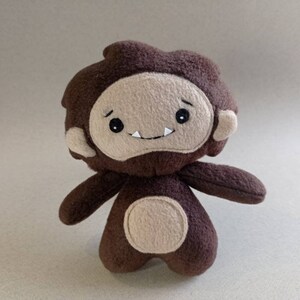 May include: A handmade plush toy in the shape of a cartoonish Bigfoot. The toy is primarily brown with a tan face, ears, and belly. It has black eyes and a small, smiling mouth. The arms and legs are short and stubby.