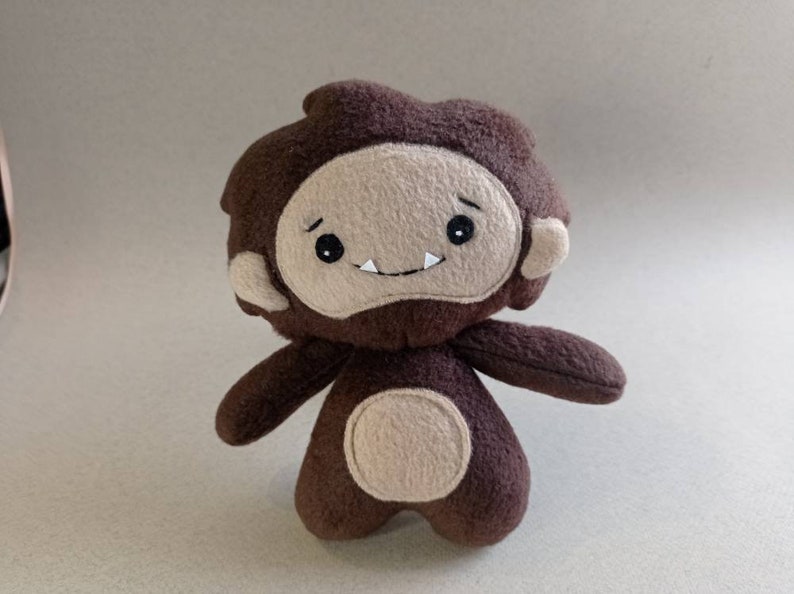 May include: A handmade plush toy in the shape of a cartoonish Bigfoot. The toy is primarily brown with a tan face, ears, and belly. It has black eyes and a small, smiling mouth. The arms and legs are short and stubby.