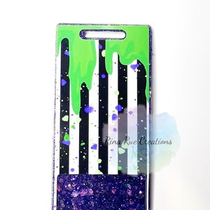 May include: A rectangular badge holder with a green top and a black and white striped background. Green slime drips down the front, with purple and green paint splatters. The bottom section is filled with purple glitter. The text "Rina Rue Creations" is visible.