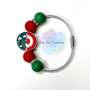 May include: A white cable bracelet with red and green beads and a charm featuring the Starbucks logo and a red target logo. The beads are accented with rhinestones.