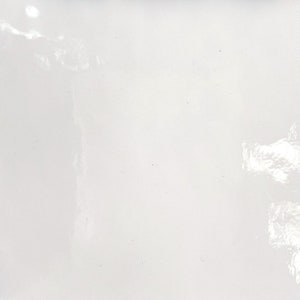 May include: A white ceramic tile with a smooth, glossy finish. The tile has a subtle, uneven texture that creates a slightly reflective surface.