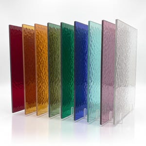 Puede incluir: Una colecci&oacute;n de nueve l&aacute;minas de vidrio rectangulares dispuestas en fila, cada una con una superficie texturizada distinta. Los colores incluyen rojo, naranja, &aacute;mbar, verde oliva, verde esmeralda, azul profundo, turquesa claro, morado y vidrio transparente.