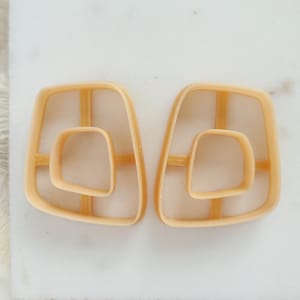 Könnte beinhalten: Ein Paar goldfarbene geometrische Ohrringe mit einem doppelschichtigen Design. Die Ohrringe sind aus einem glatten, kunststoffartigen Material gefertigt.