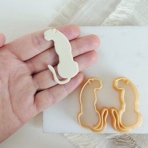 Puede incluir: Un cortador de galletas de plástico blanco con forma de guepardo sentado. Un segundo cortador de galletas, hecho de plástico marrón claro, tiene forma de dos guepardos que se miran.