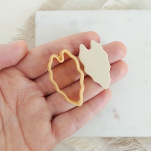Könnte beinhalten: Ein hellbeige blattförmiger Ausstecher und ein passendes blattförmiges Tonstück werden gezeigt. Der Ausstecher ist hellbraun und der Ton ist cremefarbenes Off-White. Die Gegenstände werden in einer Hand gehalten.