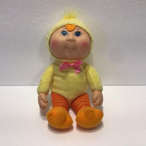 May include: A yellow plush Cabbage Patch Kid doll wearing a yellow chick costume with orange and white striped leggings and a pink bow.