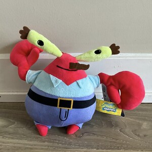 May include: A Mr. Krabs plush toy from SpongeBob SquarePants. The toy has a red head and claws, a light blue shirt, and a purple body with a black belt. It has green eyes and a brown moustache. A tag is attached to the toy.