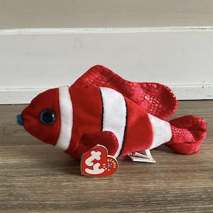 May include: A plush toy clownfish with red and white stripes, large blue eyes, and a small blue mouth. The fish has a red tail and fins with a textured pattern. A red heart-shaped tag is attached.
