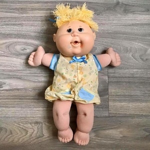Puede incluir: Una muñeca Cabbage Patch Kid con cabello rubio que lleva un vestido de estampado floral amarillo y azul. La muñeca tiene una gran sonrisa y está tumbada sobre una superficie de madera.