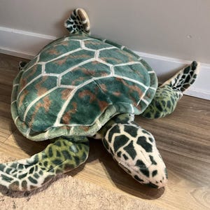 May include: A plush sea turtle toy featuring a soft, green and brown patterned shell with white outlines. The turtle has four flippers with dark green and black spots, and a head with a matching spotted pattern on a light-coloured base.