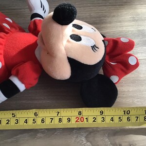 May include: A red and white polka dot plush toy of Minnie Mouse. The toy is lying on a wooden surface with a measuring tape beside it.