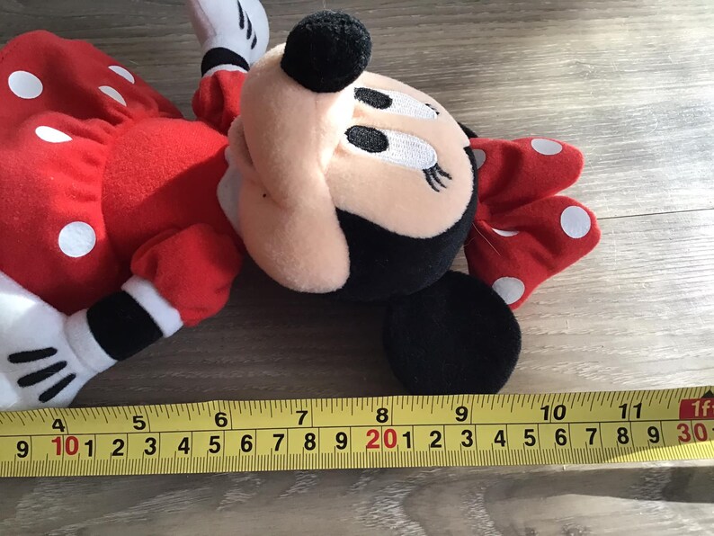 May include: A red and white polka dot plush toy of Minnie Mouse. The toy is lying on a wooden surface with a measuring tape beside it.