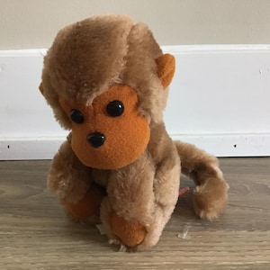 May include: A plush monkey toy with brown fur and an orange face, ears, and feet. The monkey has large black eyes and is sitting on a wooden floor. The toy is designed to resemble a monkey.