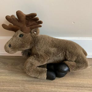 May include: A plush moose toy with soft brown fur, large dark brown antlers, and smooth black hooves. The stuffed animal is depicted in a resting position with its legs tucked underneath its body, featuring dark eyes and a small tuft of fur under its chin.