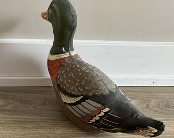 Puede incluir: Figura decorativa de pato con un diseño detallado. El pato presenta una cabeza verde, un pico marrón y un cuerpo con detalles grises, rojos y blancos. La figura está colocada sobre una superficie de madera.