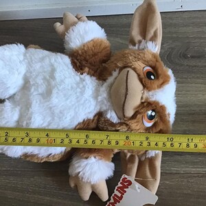 May include: A plush Gremlin toy, featuring brown, white, and tan fur. The toy is approximately 13 inches long, measured with a yellow tape measure. The Gremlin has large ears, orange eyes, and a tan snout. A tag with the word "GREMLINS" is attached.