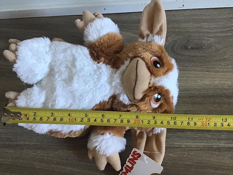 May include: A plush Gremlin toy, featuring brown, white, and tan fur. The toy is approximately 13 inches long, measured with a yellow tape measure. The Gremlin has large ears, orange eyes, and a tan snout. A tag with the word "GREMLINS" is attached.