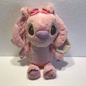 May include: Pink plush toy with large ears, a pink bow, and purple accents. The toy has brown eyes and a stitched smile.