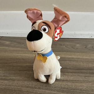 Op de afbeelding: Een pluche hondenspeelgoed met bruin en wit haar, grote flaporen en een blauwe halsband met een gele botvormige tag. De hond heeft een zwarte neus en een rood label aan zijn oor.