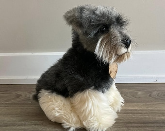 May include: A plush Schnauzer dog toy with a black and gray body, white chest and paws, and a tan tag around its neck. The toy is sitting on a wooden surface.