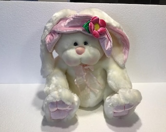 May include: White plush bunny rabbit wearing a pink hat with a pink flower. The bunny has pink feet and a pink bow around its neck.