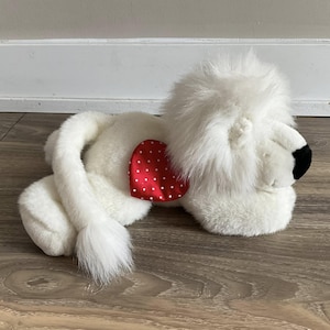 May include: White plush lion with a red heart-shaped bow with white polka dots. The lion is lying on its side with its head resting on the floor.