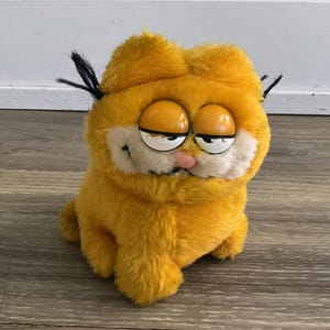 Könnte beinhalten: Ein Plüschtier in Form einer Cartoon-Katze mit leuchtend orangefarbenem Fell, einer weißen Schnauze und einer kleinen rosa Nase. Die Figur hat große, halb geschlossene Kunststoffaugen mit schwarzen Pupillen, ein dezentes Grinsen und dünne schwarze Schnurrhaare.