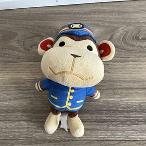Pode incluir: Um brinquedo de peluche de macaco vestindo um uniforme azul e amarelo com um chapéu vermelho e amarelo. O macaco tem um nariz castanho e olhos castanhos.
