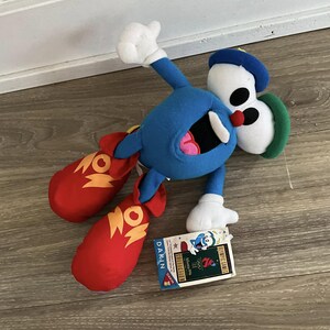 May include: A blue plush toy with red and yellow boots. The toy is wearing a green and blue hat and has large eyes. The toy is labeled "Olympic Games Atlanta 1996" and "Dakin Collection".