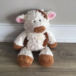 Pu&ograve; includere: Un giocattolo di peluche di mucca bianco e marrone con orecchie rosa. La mucca &egrave; seduta con le gambe incrociate e ha una consistenza soffice.
