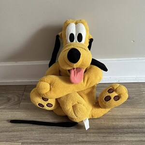 Pode incluir: Um brinquedo de pelúcia do Pluto, um cão amarelo com orelhas pretas e uma cauda preta. O brinquedo tem uma língua rosa saindo e manchas marrons nas patas. O brinquedo está sentado em um chão de madeira.