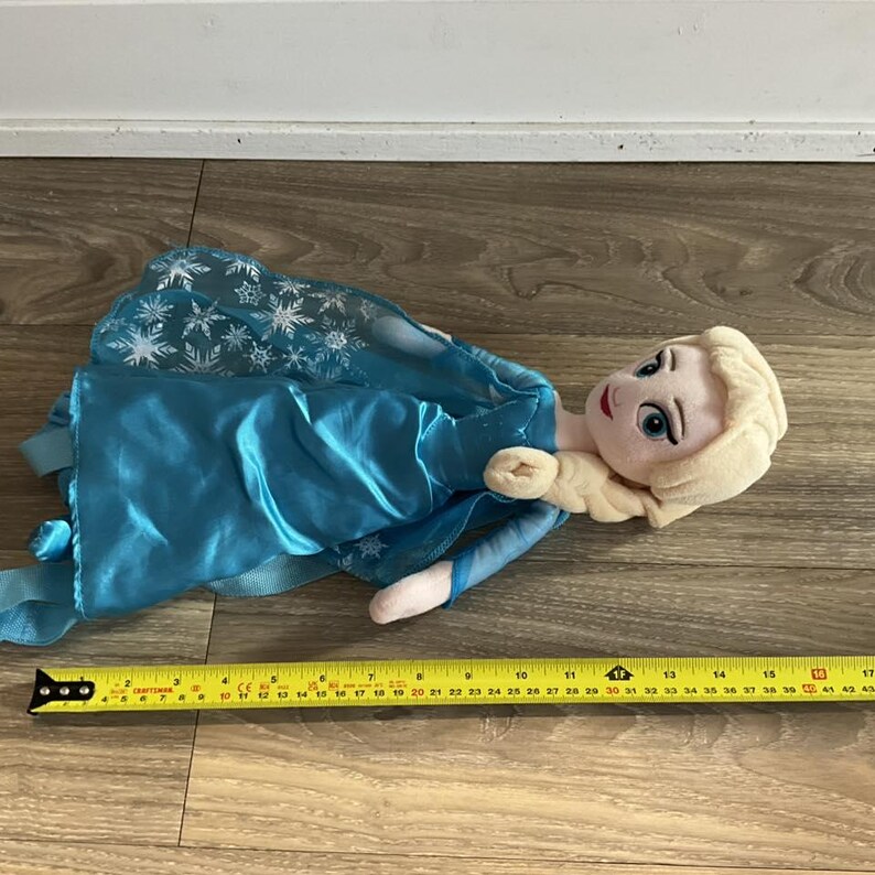 May include: A plush doll of a character from the movie Frozen, wearing a blue dress with a snowflake design on the cape. The doll has blonde hair and is lying on a wooden surface. A yellow measuring tape is next to the doll.