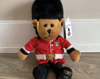 May include: A plush toy bear dressed as a British guardsman. The bear is wearing a red uniform with a black hat, a white belt, and a gold button. The bear is sitting with its legs crossed and has a tag that says "Guardsman Bear" on it. The bear's feet have the words "Tower of London" on them.