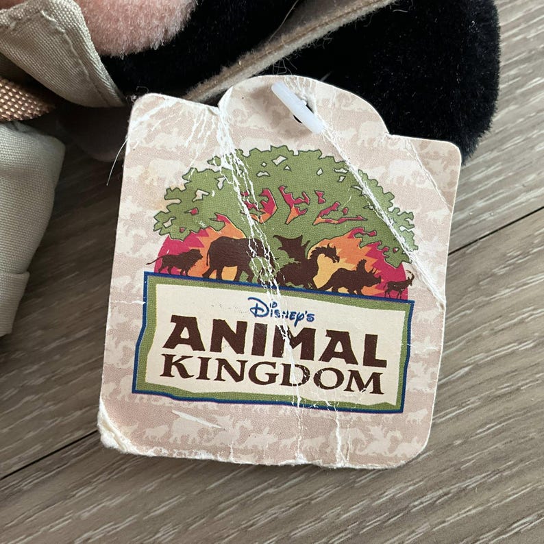 May include: A square-shaped paper hang tag with rounded corners featuring a graphic of a green tree and various animal silhouettes including a lion, elephant, dragon, and rhinoceros. The text reads Disney's ANIMAL KINGDOM in dark brown letters.
