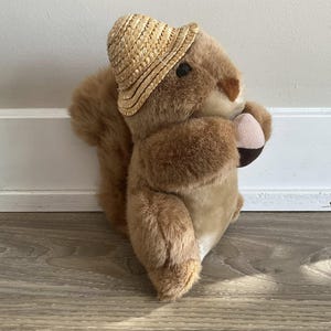 May include: A brown plush squirrel wearing a straw hat. The squirrel is holding a small brown object in its paws.