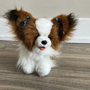 Op de afbeelding: Een pluche Papillon-hondenspeelgoed met bruin en wit haar, grote oren en een kleine roze tong. Het speelgoed heeft blauwe strikjes in de oren en een zwarte neus. De hond zit op een houten oppervlak.