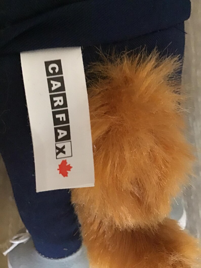 Pu&ograve; includere: Un'etichetta bianca con la parola "CARFAX" in maiuscolo nero, con un logo a forma di foglia d'acero rossa. L'etichetta &egrave; attaccata a un peluche con pelliccia arancione e tessuto blu.