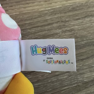 K&ouml;nnte beinhalten: Ein wei&szlig;es Etikett mit dem Text "HugMees Original by Squishmallows TM" in bunten Buchstaben. Das Etikett ist an einem rosa-wei&szlig;en Pl&uuml;schtier mit gelben Akzenten befestigt.