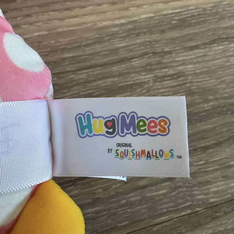 K&ouml;nnte beinhalten: Ein wei&szlig;es Etikett mit dem Text "HugMees Original by Squishmallows TM" in bunten Buchstaben. Das Etikett ist an einem rosa-wei&szlig;en Pl&uuml;schtier mit gelben Akzenten befestigt.