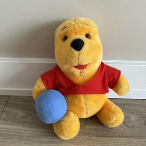 Könnte beinhalten: Ein gelbes Winnie-Puuh-Plüschtier, das sitzt, ein rotes Oberteil trägt und einen blauen Ball hält. Das Spielzeug hat eine schwarze Nase und einen roten, lächelnden Mund. Der Hintergrund ist eine weiße Wand und ein Holzboden.