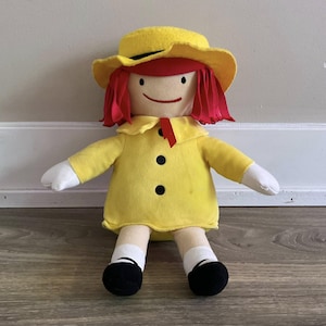 May include: A handmade doll with a yellow hat and dress, red hair, and black shoes. The doll has a simple, smiling face and white gloves. The dress has two black buttons and a red ribbon. A charming, whimsical toy.
