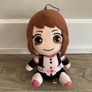 May include: A plush doll of a character with brown hair and large brown eyes. The doll wears a black and pink outfit with a white belt and pink accents. The doll is sitting with its legs extended and has a black string attached to the top of its head.