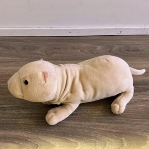 May include: A beige plush naked mole rat toy. The soft toy has a wrinkled body, small black eyes, and pink ears. It is lying on a wooden surface.